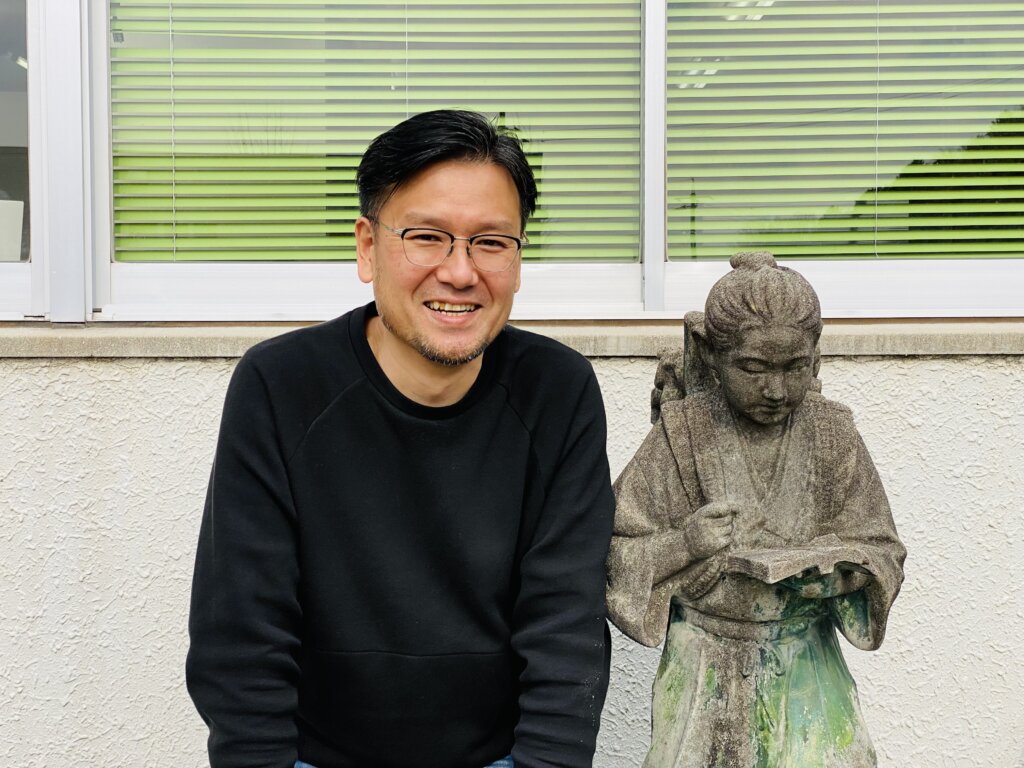 二宮金次郎像の横でインタビューに答える代表取締役の井掛 雅祥さん